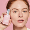 A young woman holds up a bottle of Saturday Skin Wide Awake Brightening Eye Cream next to her face, with several dollops of it applied beneath her eye