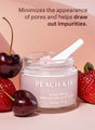 A jar of Peach & Lily Pore Proof Perfecting Clay Mask with a small spoon stuck in the top and surrounded by cherries and strawberries, which matches the color of the clay mask, and a highlight of how the product minimizes the appearance of pores and helps draw out impurities